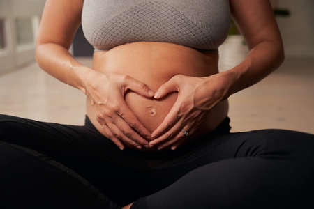 Caucasian pregnant mother sitting on yoga mat forming a heart shape with her hands on bellyの写真素材