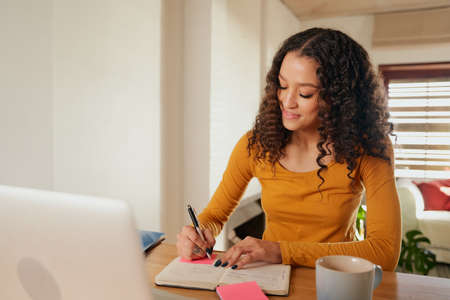Happy Multi-cultural female focussed, writing in notebook. Young professional remote working with laptop in modern apartmentの写真素材