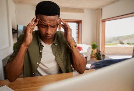 Exhausted Young adult black male massaging his temples while he works on laptop from home in modern kitchen.の写真素材