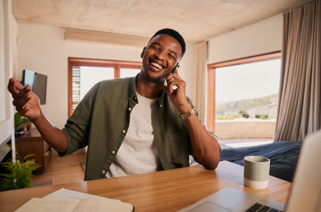 Young adult black male smiling after receiving good news on a phone call. Working from laptop at home in modern apartment.の写真素材