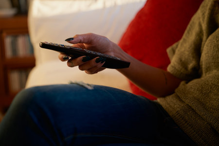 Hand of Multicultural young adult female using remote control for TV. Modern apartment, sitting on...