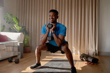 African American male squatting in lounge holding dumbbell weight, working out at homeの写真素材