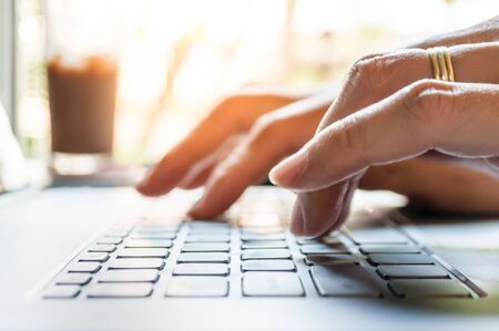 Asian man hands typing on laptop computer with blurred coffee glass and view outside window.