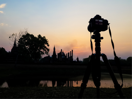 Silhouette of camera on tripod and ancient ruin in Thailand against sunset sky. The temple named Wat Mahathat, part of Sukhothai historical park, ancient kingdom in thirteenth centuries.の写真素材