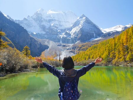 Young woman raising her arms up in the air in Pearl Lake or Zhuoma La Lake with snow mountain in autumn in Yading Nature reserveの写真素材