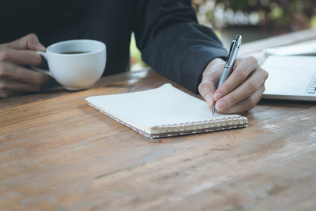 Close up businessman write notebook and and holding coffee cup on wood table.の写真素材