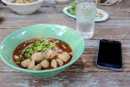 Thai noodle in green Bowl , Black display Smart phone beside bowl noodles.All placed on wooden...