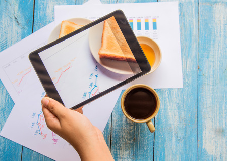 Tablet in hand , Toast , Honey , Coffee cup , Graph data analysis placed on blue wooden floor.Graph...