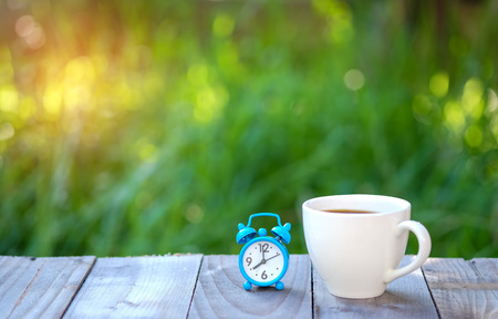 Clock and coffee cup on wooden with green blurred background.