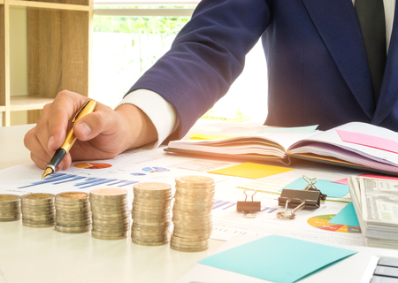 Businessman has a pen in his right hand analysis of graph data,On the desk there are coins, dollars,...