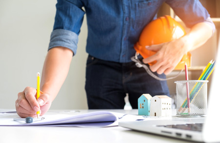 Architects are using drawing pencils,On the other hand holds a safety hat,On the writing desk there are model houses, laptop and house plans.の写真素材