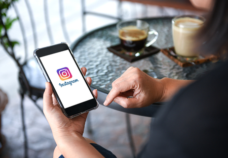 CHIANGMAI, THAILAND - August 26, 2018. Instagram
 application on smart phone display in hand with coffee on table background in cafe.のeditorial素材