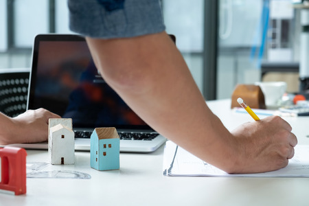 Architects are writing home plan and using laptop.Model house on the table.(Taken from the back)の写真素材