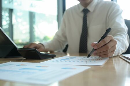 Young businessman is checking data on a graph and using a laptop on an office desk.He uses the pen...