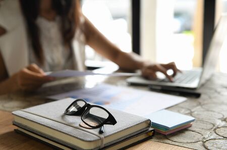 Glasses placed on notebooks, background blurred people are sitting using laptop.