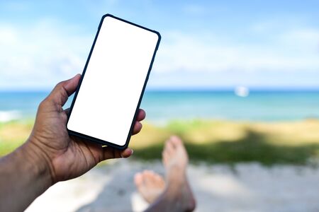 Men hand holding a white screen blank phone.He slept on the beach with sea background.の写真素材