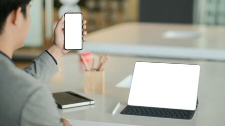 Young businessman hold a blank screen smartphone in hand and a blank screen laptop on the desk.の写真素材