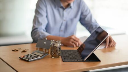 Cropped shot of a Laptop, Coin in a glass bottle, Calculator with a model house on the table and a background blurred people sitting watching documents.の写真素材
