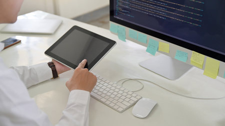 A programmer men using a tablet in his hand with a computer screen on his desk.の写真素材