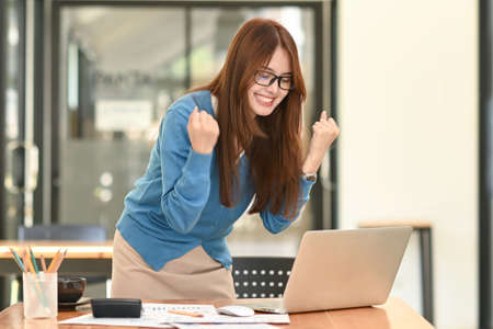 Happy businesswoman raising her hand on investment success, looking at laptop on the office desk smiling face, reaching a business deal, profitable business.の写真素材