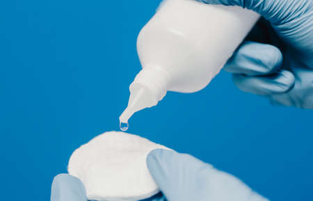 A guy in a medical glove holds cotton pads and a second-hand bottle. Blue background. Medical and cosmetology contentの写真素材