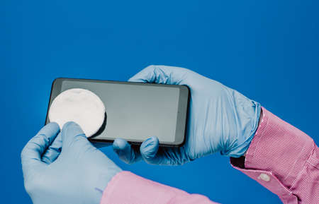 A guy in a medical glove wipes his phone with cotton pads. Blue background. Antibacterial effect, disinfects, disinfectsの写真素材