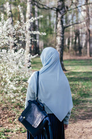 Muslim woman in hijab in a park next to a blossoming tree. Blue shawl and blue backpack. Modern muslim woman. Spring Islam. Islamic woman. Education in Islam. Muslim studentの写真素材
