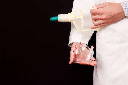 Jars for hijama, bloodletting and a pump in the hands of a doctor in a white coat on a black...