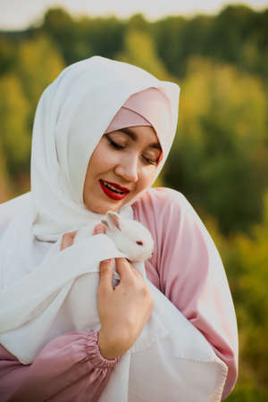 Muslim woman hold a small white rabbit in her arms. A portrait of a Muslim woman in a pink dress and a scarf, a pearl bracelet. Tenderness.の写真素材