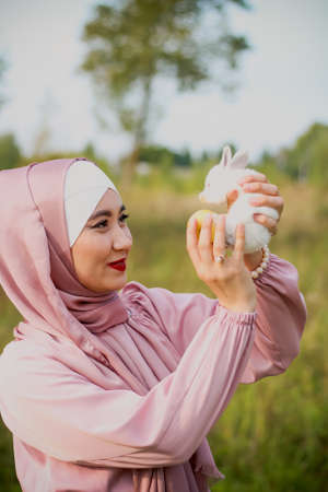 Muslim woman hold a small white rabbit in her arms. A portrait of a Muslim woman in a pink dress and a scarf, a pearl bracelet. Tenderness.の写真素材