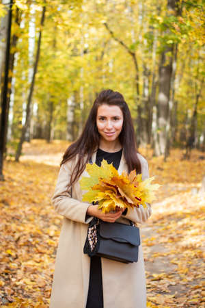 Brunette woman in autumn park and autumn leaves. Small black bag. Autumn mood. Yellow, red and green leaves. Beige coat.の写真素材