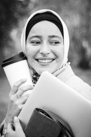 A Muslim woman in a hijab in a beige trench coat stands with a cup of coffee and a laptop in the park. Muslim business woman. Student. Close plan, face. Black and white photographyの写真素材