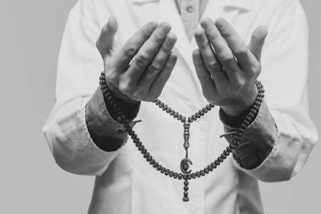 A young man in a white robe raised his hands up and prayed with a Muslim rosary. Medical gloves. Doctor praying. A Muslim makes dua. Black and white photo.の写真素材