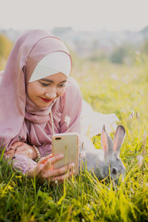 A Muslim woman in a hijab is photographed on the phone with a rabbit. Pink scarf. Grass field