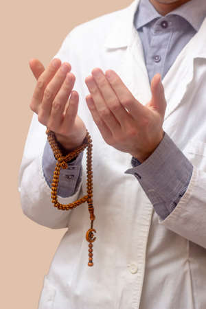 A young man in a white robe raised his hands up and prayed with a Muslim rosary. Beige background. Doctor praying. A Muslim makes dua.の写真素材
