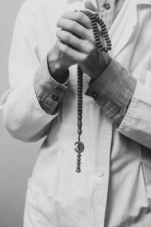 A young man in a white robe raised his hands up and prayed with a Muslim rosary. Medical gloves. Doctor praying. A Muslim makes dua. Black and white photo.の写真素材