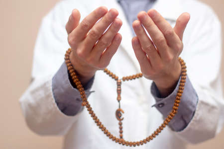 A young man in a white robe raised his hands up and prayed with a Muslim rosary. Beige background. Doctor praying. A Muslim makes dua.の写真素材