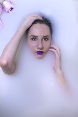Young beautiful woman with bright violet make-up in milk bath with violet and white flowers. Healthy...