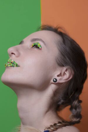 Young woman portrait with a candy makeup - multicoloured pastry topping pearls on her lips and eyelids. On a green and orange background. Easter theme.の写真素材