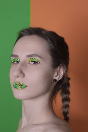 Young woman portrait with a candy makeup - multicoloured pastry topping pearls on her lips and eyelids. On a green and orange background. Easter theme.の写真素材