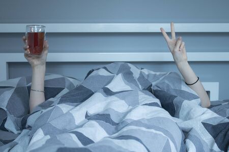 Hands of a girl from under a blanket holding a cup of tea with a double bottom, showing a pease sign...