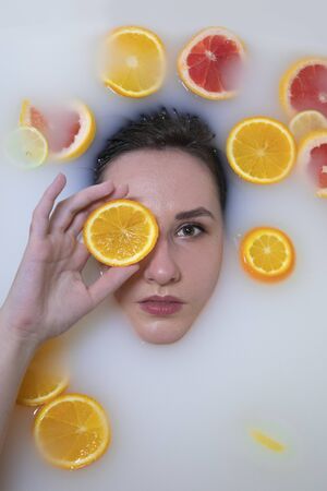 Woman portrait in milk bath with oranges, lemons and grapefruits. Healthy dewy skin. Fashion model girl, spa and skin care concept. Spring colours - yellow, orange, red.の写真素材