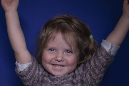 portrait of happy a three year old blond girl with blue eyes on a blue background - hands up