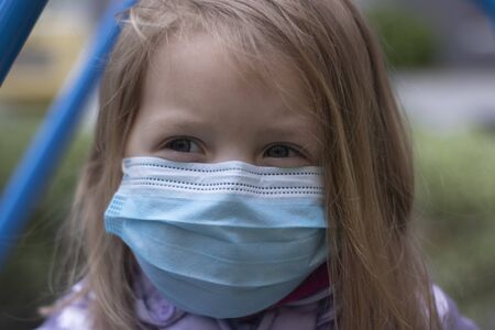 Portrait of a cute blonde little girl wearing a Medical Disposable Face Mask, playing on a...
