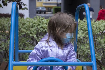 Portrait of a cute blonde little girl wearing a Medical Disposable Face Mask, playing on a playground during quarantine. Stay at home. Isolation. Coronavirus and Air pollution concept.の写真素材