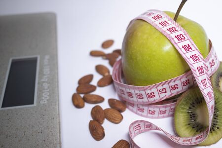 Red measuring tape, apple, kiwi and almonds near bathroom scales. Diet and Healthy life, loss weight, sport concept. Copy space. Isolated. White backgroundの写真素材