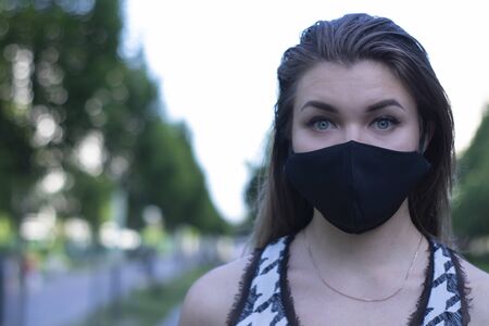 Pretty young blonde woman in medical black face mask. Wearing a t-shirt and jeans shorts. in a park. modern reality. covid-19 concept. copy space. High quality photoの写真素材