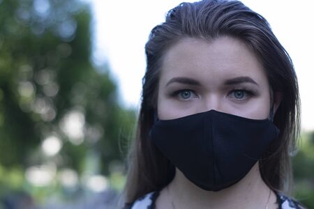 Pretty young blonde woman in medical black face mask. Wearing a t-shirt and jeans shorts. in a park. modern reality. covid-19 concept. copy space. High quality photoの写真素材
