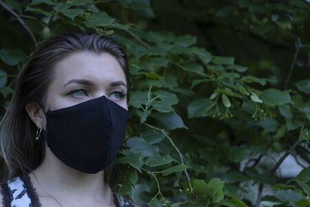 Pretty young blonde woman in medical black face mask. in a bush, forest. covid-19 concept. modern...