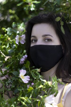 Attractive young brunette woman in yellow t-shirt and medical black face mask. in bush with pink flowers. covid-19 concept. modern reality. copy space. High quality photoの写真素材